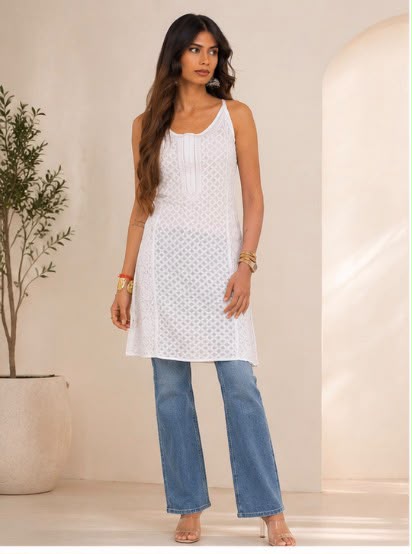 A woman modeling a white sleeveless chikankari short kurti  with blue wide-leg jeans, standing in a warm-toned indoor setting with a potted plant on the left.