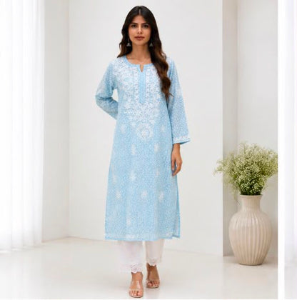 A woman modeling a light blue chikankari embroidered kurta with white pants in a bright, minimalist room with a decorative vase of flowers.
