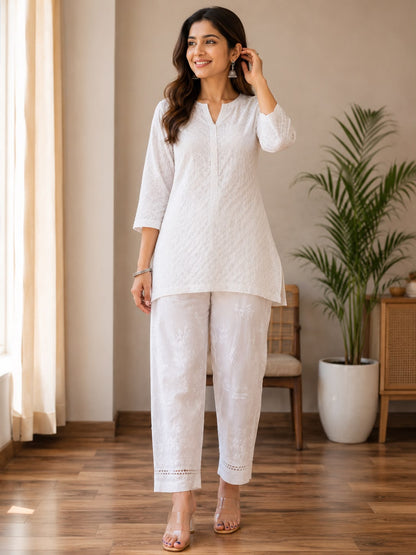 A woman modeling a white textured kurta and  chikankari trouser in a bright indoor room with wooden floors, a potted plant, and neutral walls.